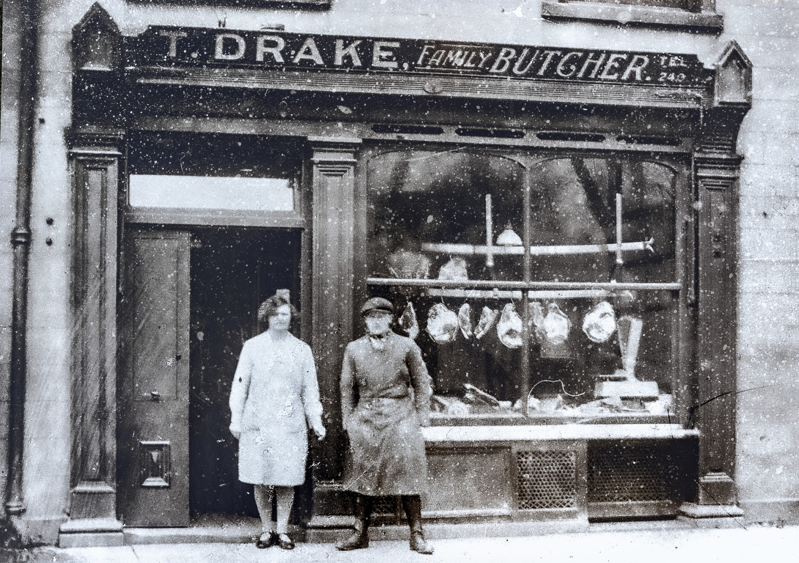 Archival photograph of the T. Drake Family Butcher shopfront on Otley Street, Skipton, early 1900s. Two figures in white aprons stand outside the door; carcasses hang in the window.